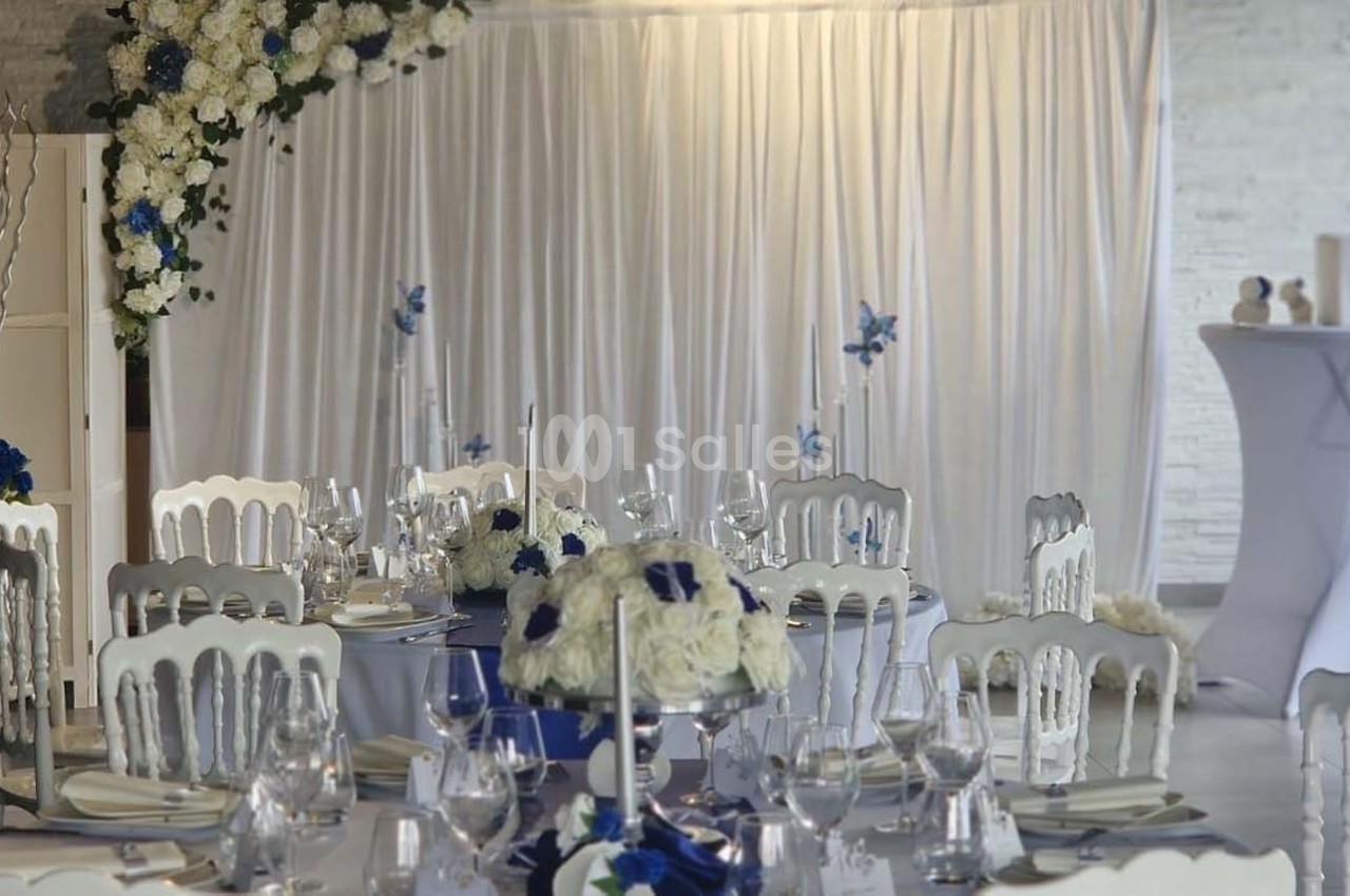Salle décorée pour un événement avec des tables dressées, des chaises blanches et des arrangements floraux bleus et blancs.