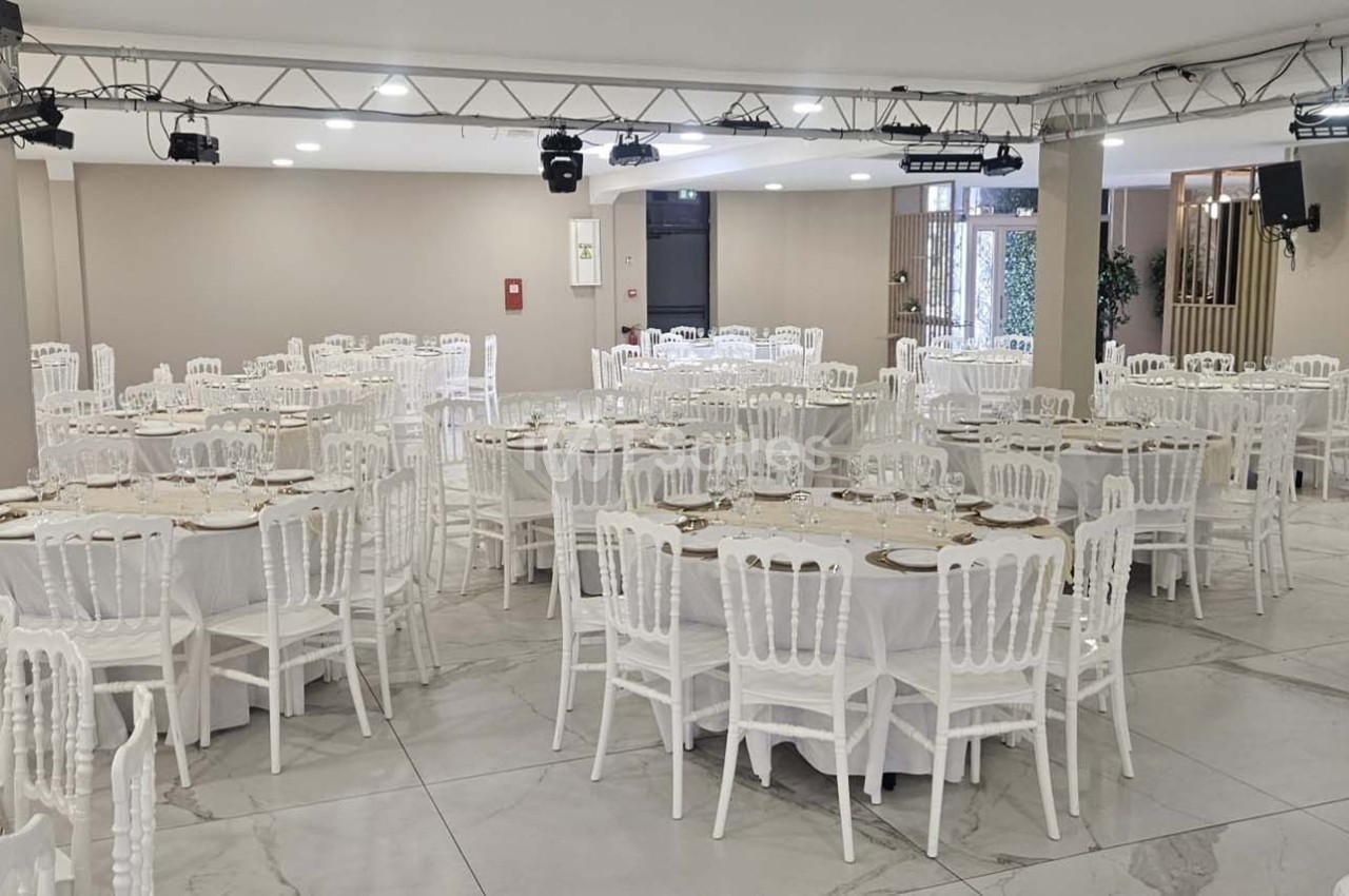 Salle de réception lumineuse avec des tables rondes dressées et des chaises blanches, prête pour un événement.
