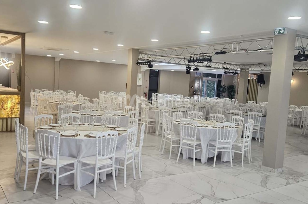 Salle de réception lumineuse avec tables rondes dressées, chaises blanches et décorations sobres.