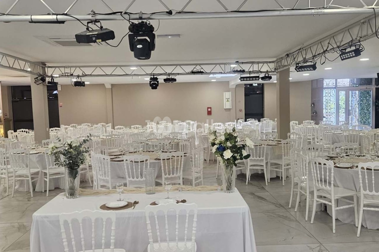 Salle de réception lumineuse avec tables rondes dressées, chaises blanches et décorations florales.