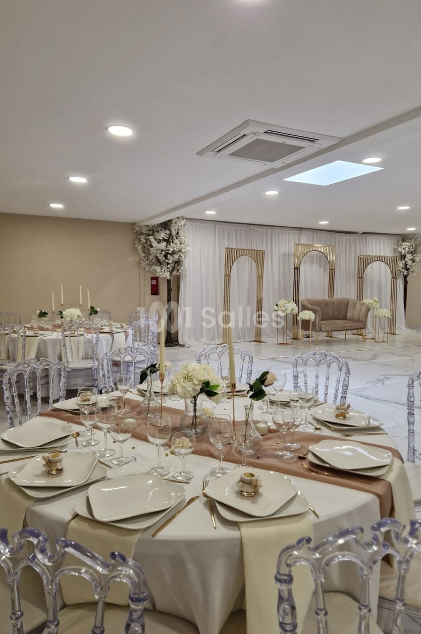 Salle de réception élégante avec tables dressées, chaises transparentes et décorations florales dans des tons neutres.