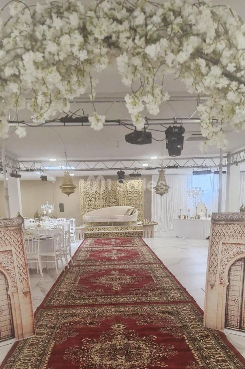 Décoration de salle de réception avec arche florale, tapis rouges ornés et estrade ornée de motifs dorés.