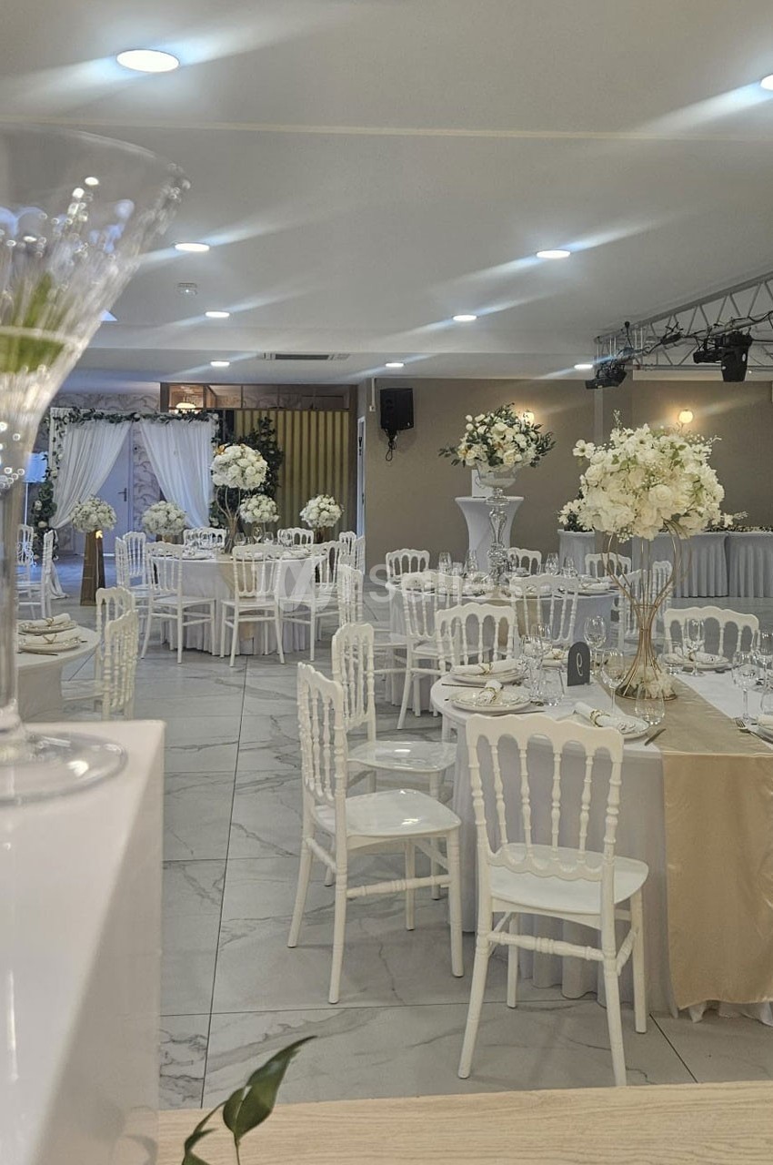 Salle de réception décorée avec des tables dressées, chaises blanches et arrangements floraux blancs.