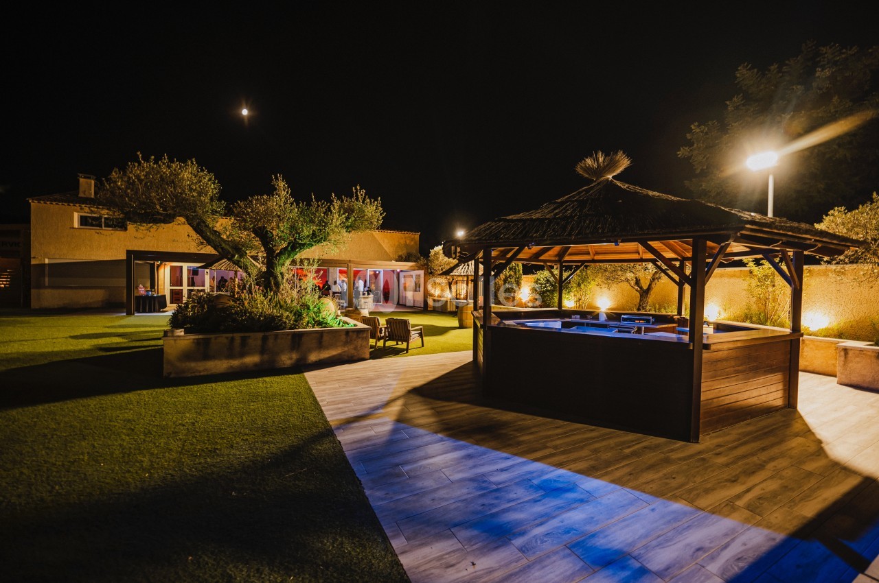 Jardin éclairé de nuit avec un jacuzzi sous une pergola en bois, un olivier et une maison en arrière-plan.