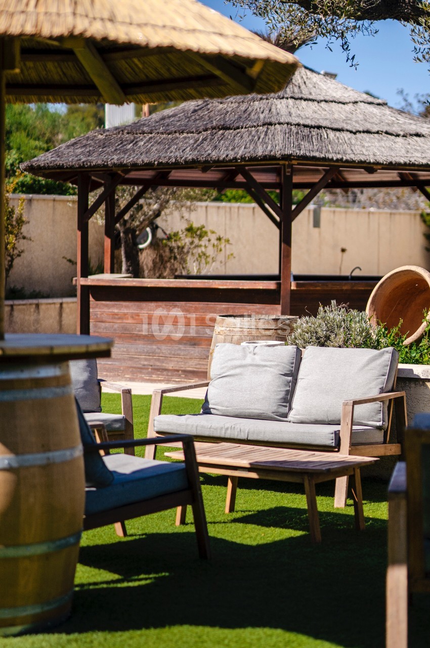 Mobilier de jardin en bois avec coussins gris, installé sur une pelouse artificielle près de pergolas en paille.