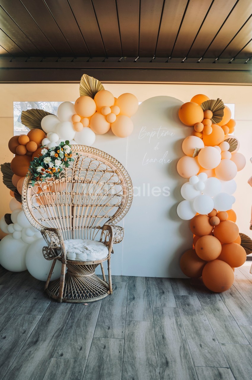 Fauteuil en rotin devant une arche décorée de ballons orange et blancs, dans un espace intérieur avec sol en bois.