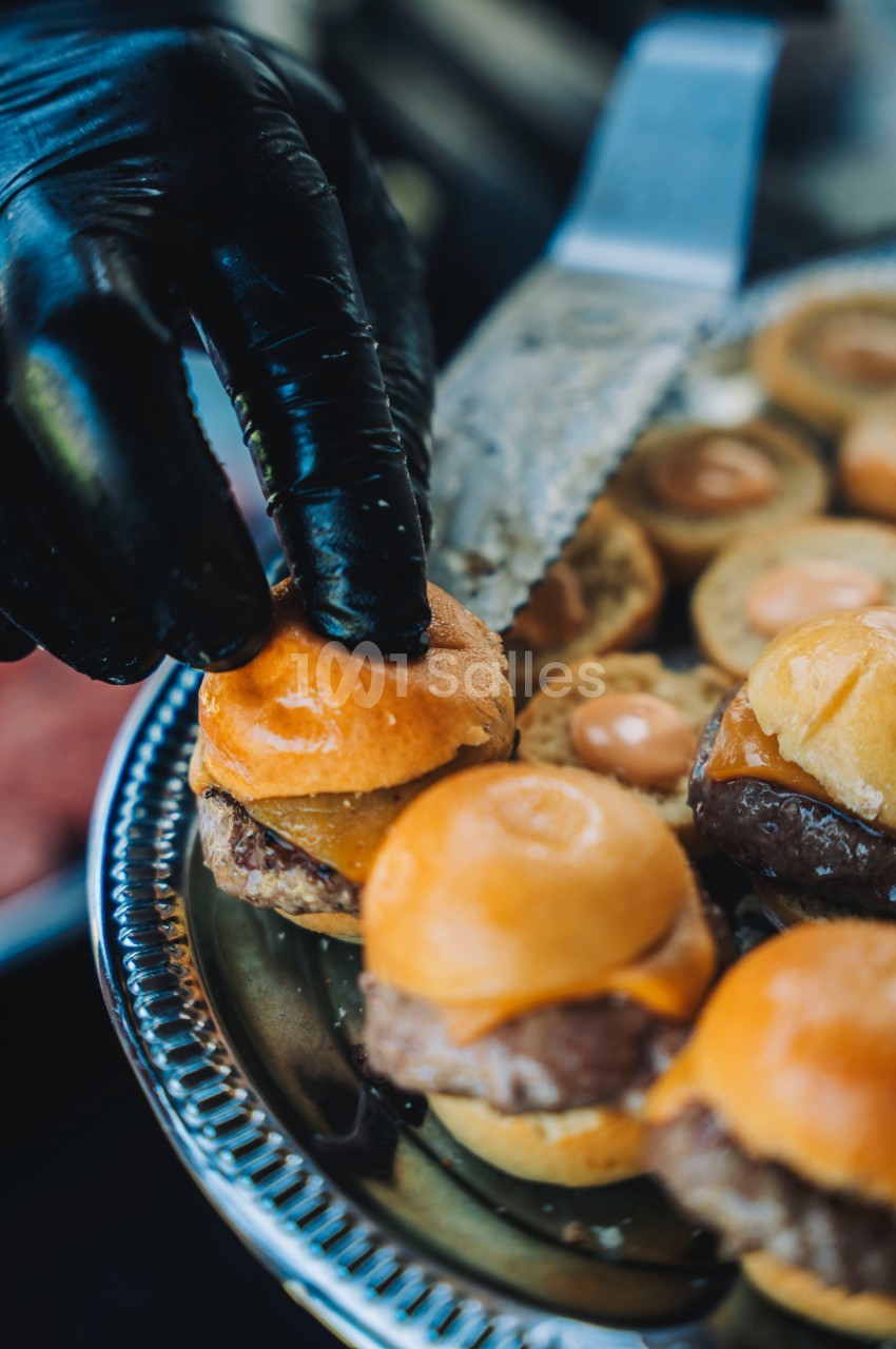 Main gantée plaçant un mini-burger sur un plateau argenté avec d'autres mini-burgers garnis de sauce.