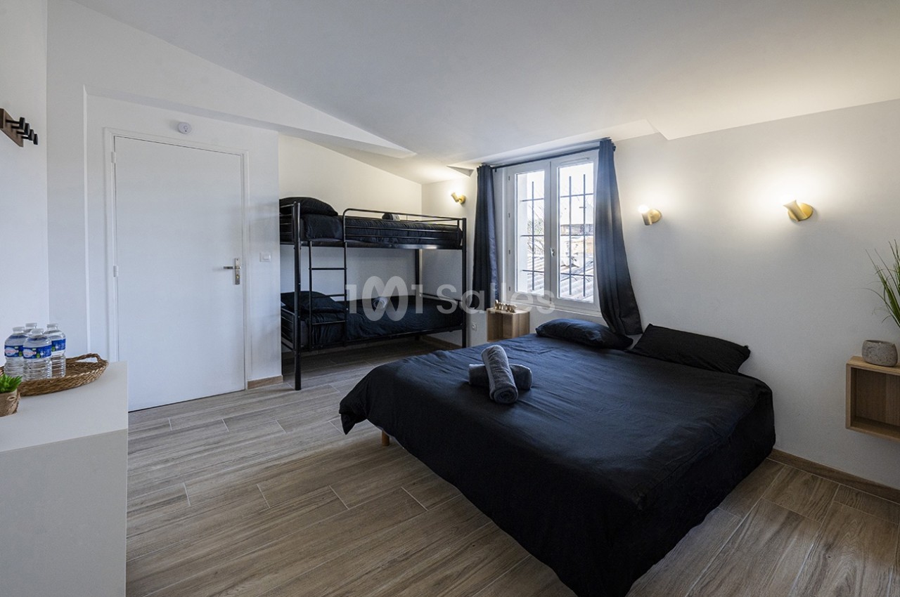 Chambre lumineuse avec un lit double, des lits superposés, sol en bois clair, murs blancs et fenêtre avec rideaux sombres.