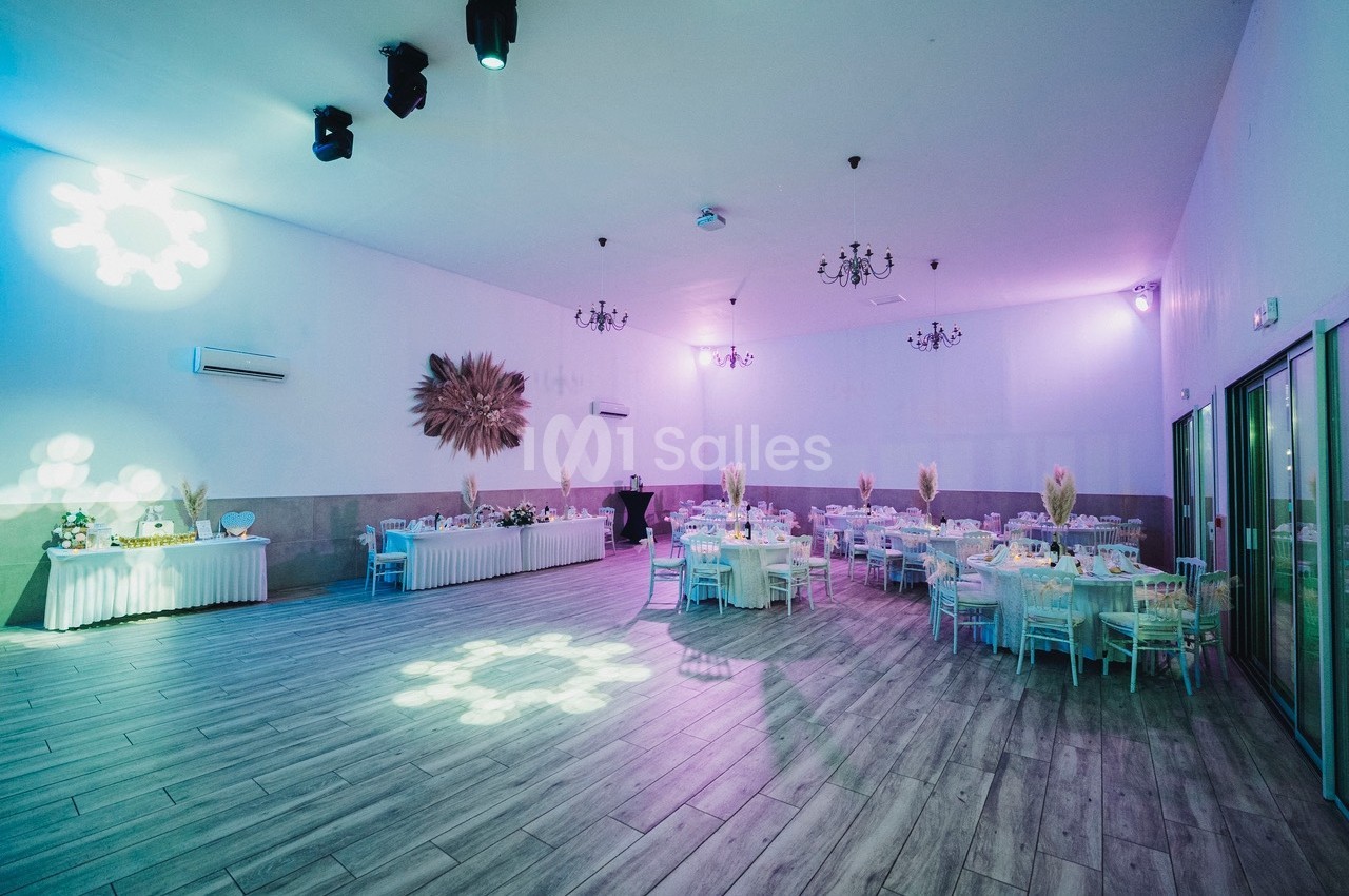 Salle de réception décorée avec des tables rondes dressées, éclairage tamisé et projecteurs lumineux au plafond.