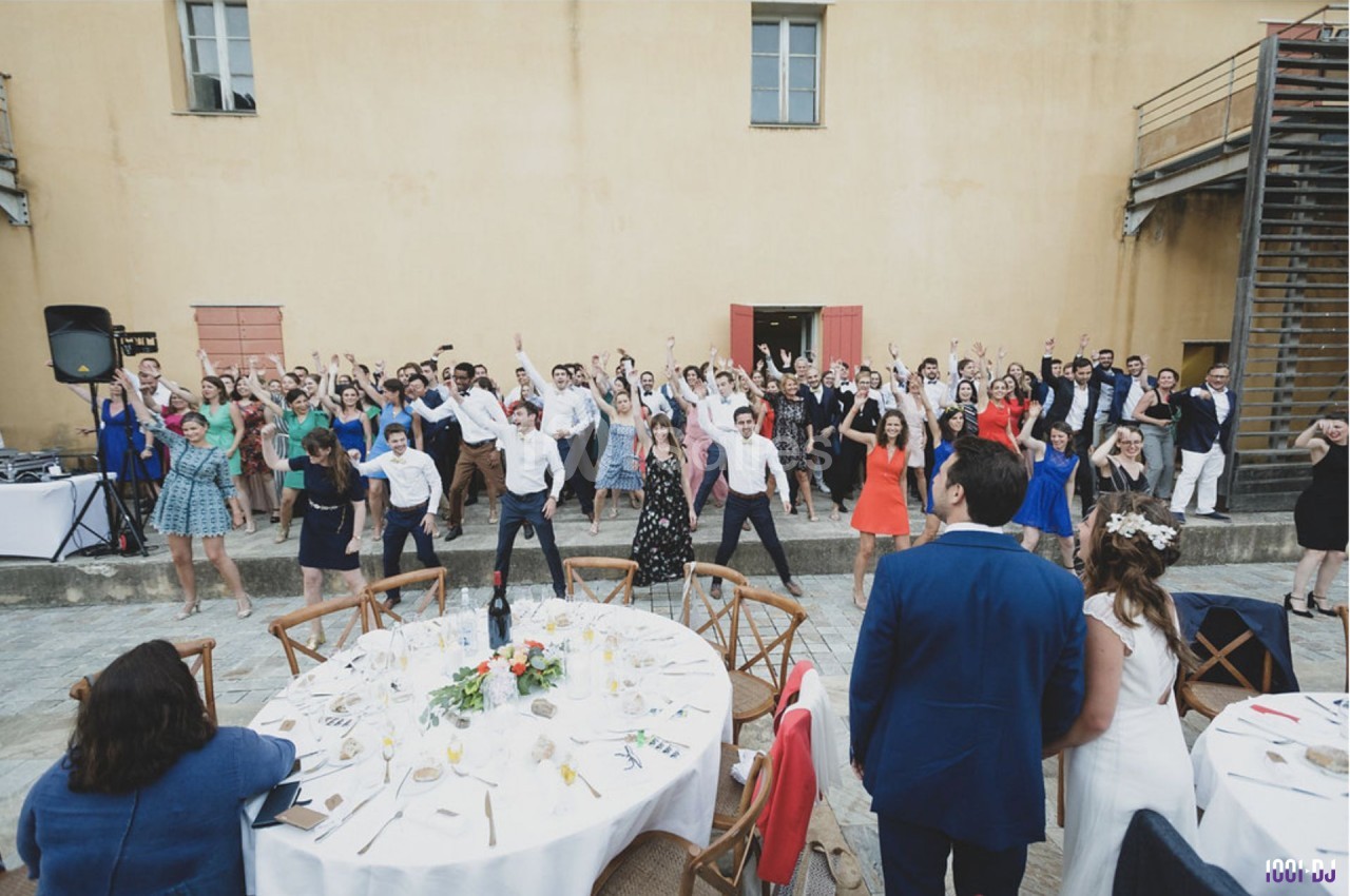 Des invités dansent en groupe lors d'une réception de mariage en extérieur devant un bâtiment beige.