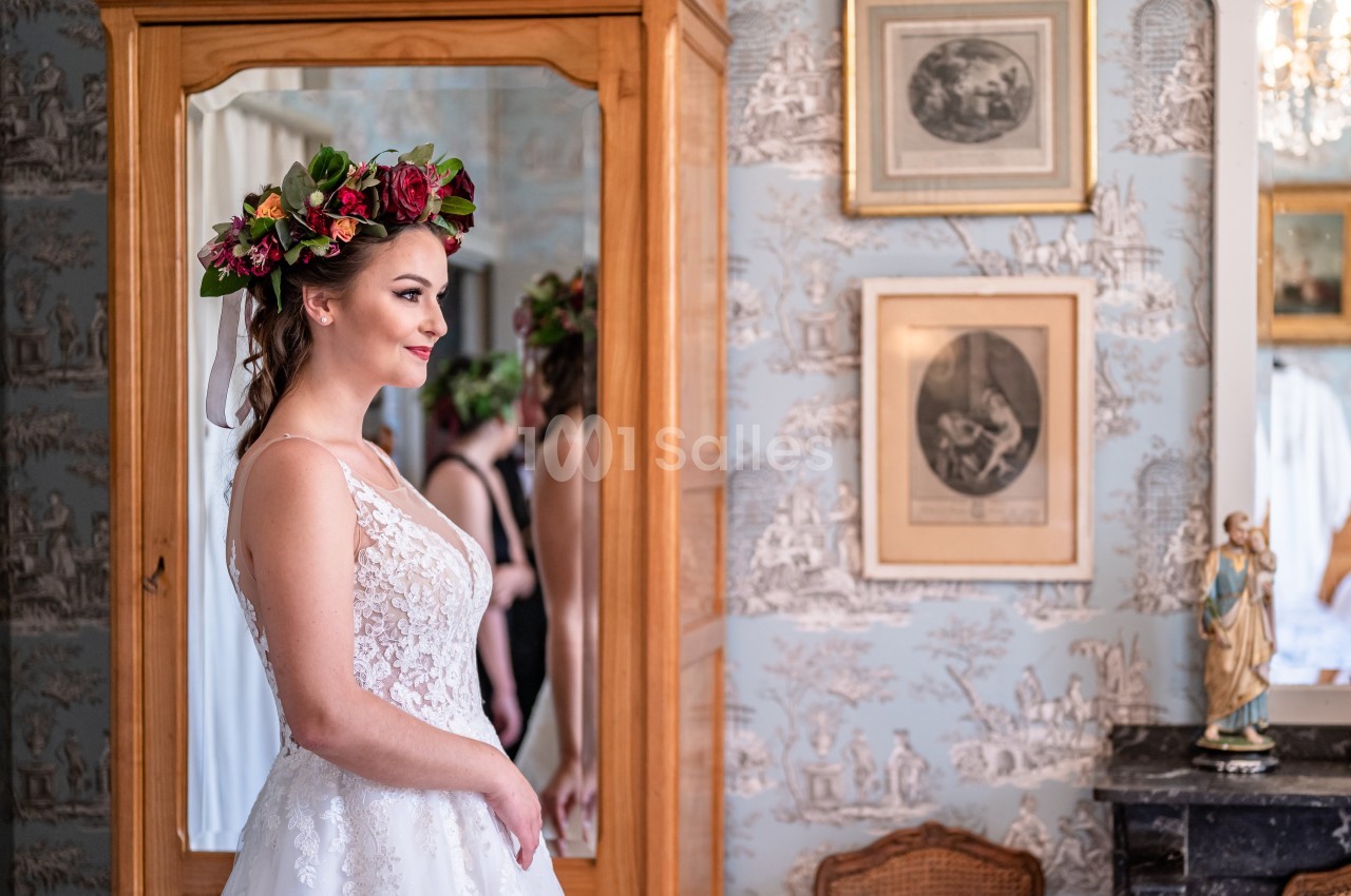 Femme en robe de mariée avec une couronne de fleurs, debout devant un miroir dans une pièce décorée de cadres et papiers…