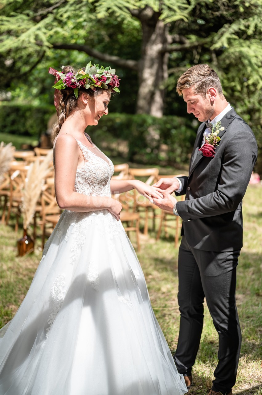 Un couple échange des alliances lors d'une cérémonie de mariage en extérieur, entouré de verdure et de chaises en bois.