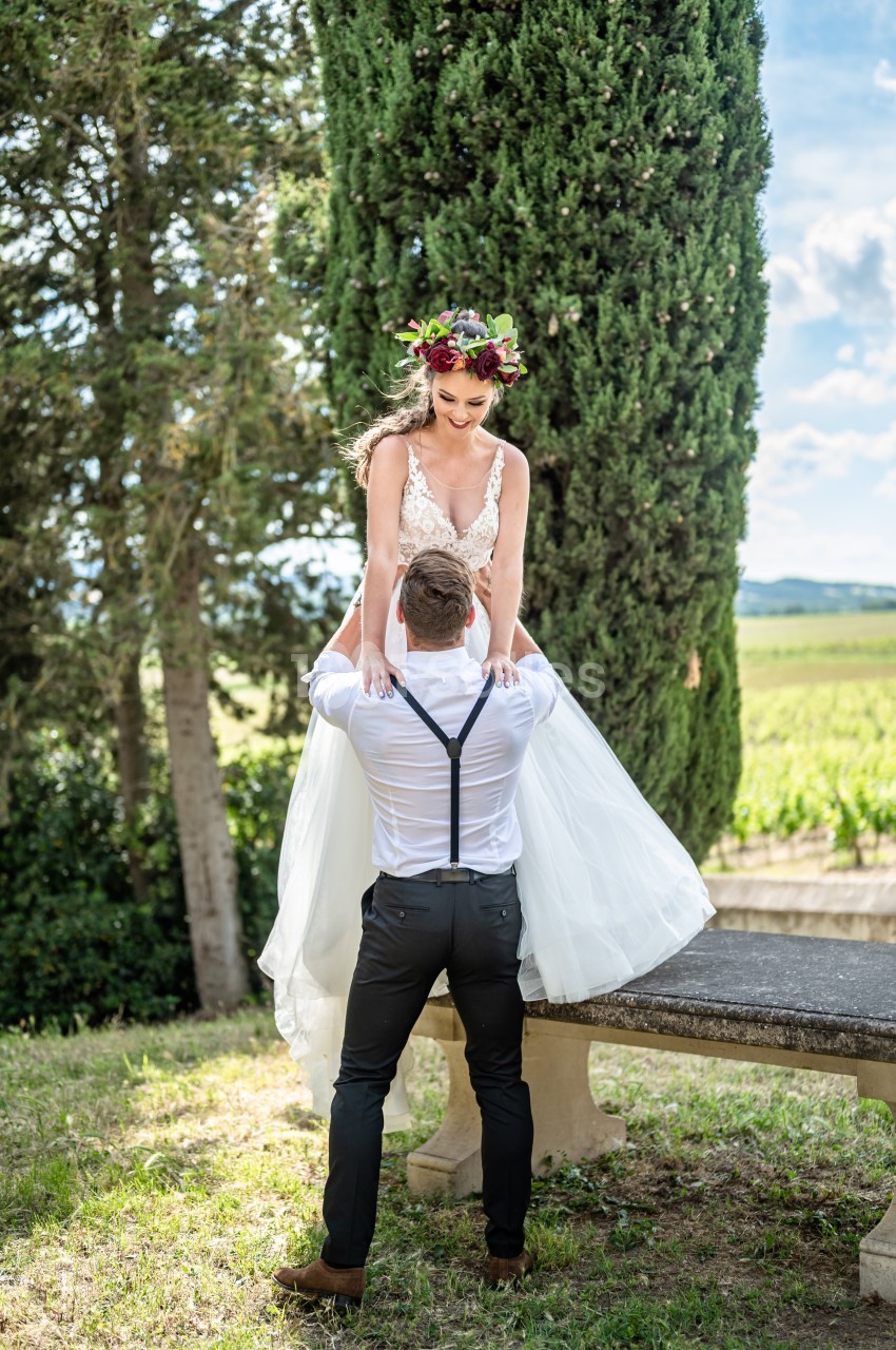 Un couple en tenue de mariage, l'homme soulève la femme devant un paysage champêtre avec des arbres et un banc en pierre.
