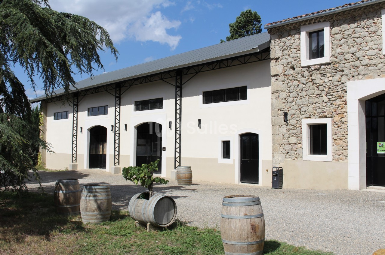 Bâtiment en pierre et crépi avec des portes noires, entouré de barriques en bois et d'arbres sous un ciel dégagé.
