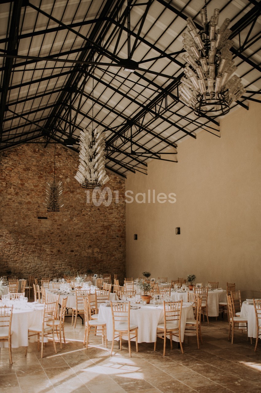 Salle de réception lumineuse avec tables rondes dressées, chaises en bois clair et lustres suspendus sous une verrière.