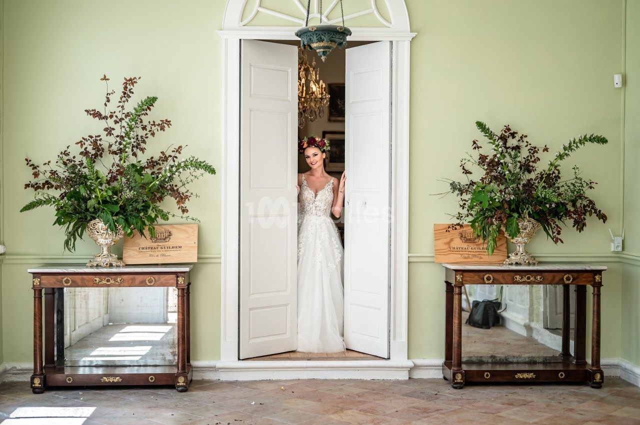 Une mariée en robe blanche se tient dans l'encadrement d'une porte blanche, entourée de décorations florales.