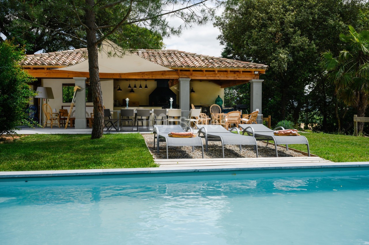 Terrasse avec mobilier en bois sous une pergola, entourée de verdure, près d'une piscine et de chaises longues.