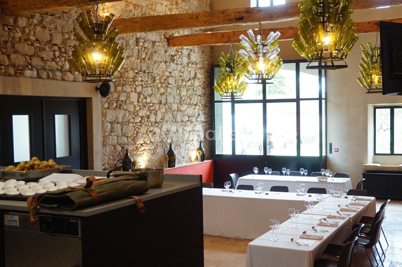 Salle de réception lumineuse avec murs en pierre, poutres apparentes, tables dressées et suspensions en verre coloré.