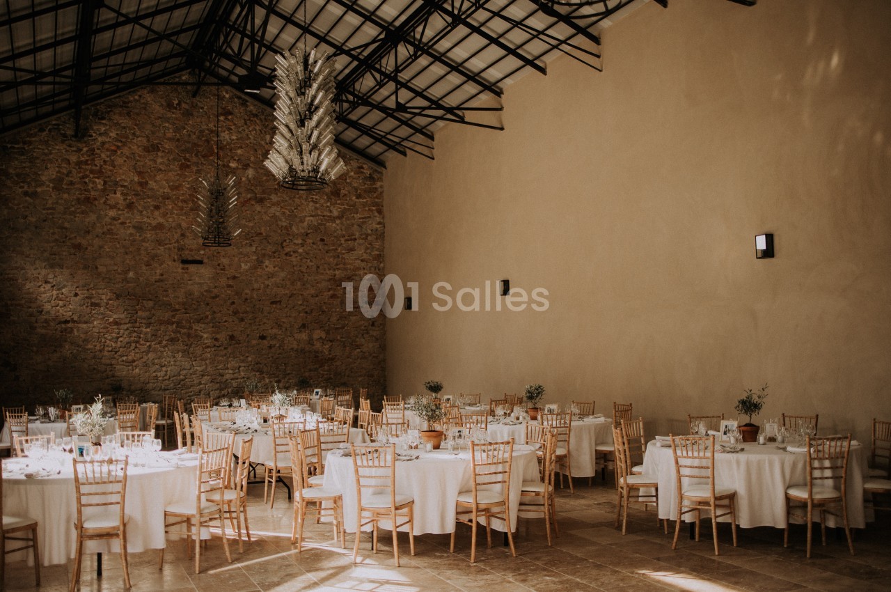 Salle de réception avec murs en pierre, tables rondes dressées et chaises en bois sous un plafond en structure métallique.