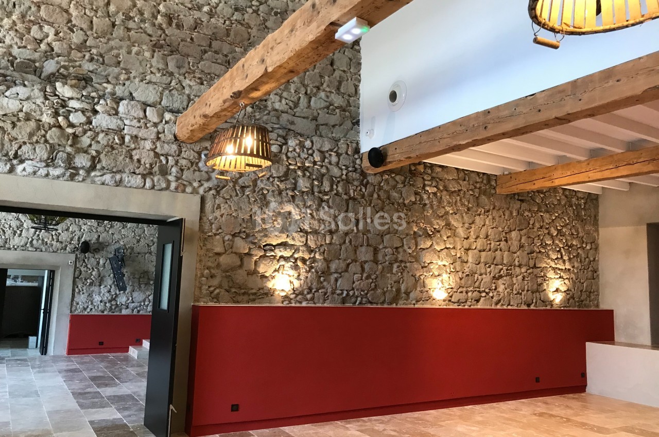 Salle avec murs en pierre apparente, poutres en bois au plafond, sol en pierre et éclairage suspendu.