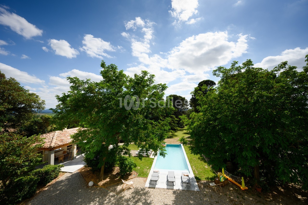 Vue aérienne d'une piscine entourée de verdure, avec des chaises longues et un bâtiment annexe sous un ciel dégagé.