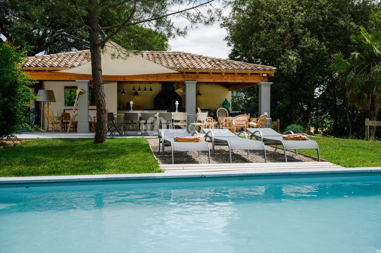Terrasse avec mobilier de jardin sous une pergola, vue sur une piscine entourée de pelouse et d'arbres.