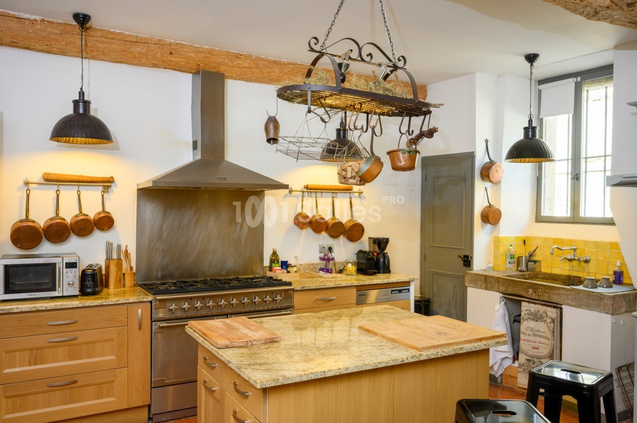 Cuisine rustique avec plan de travail en marbre, cuisinière en inox, casseroles en cuivre accrochées et éclairage suspendu.