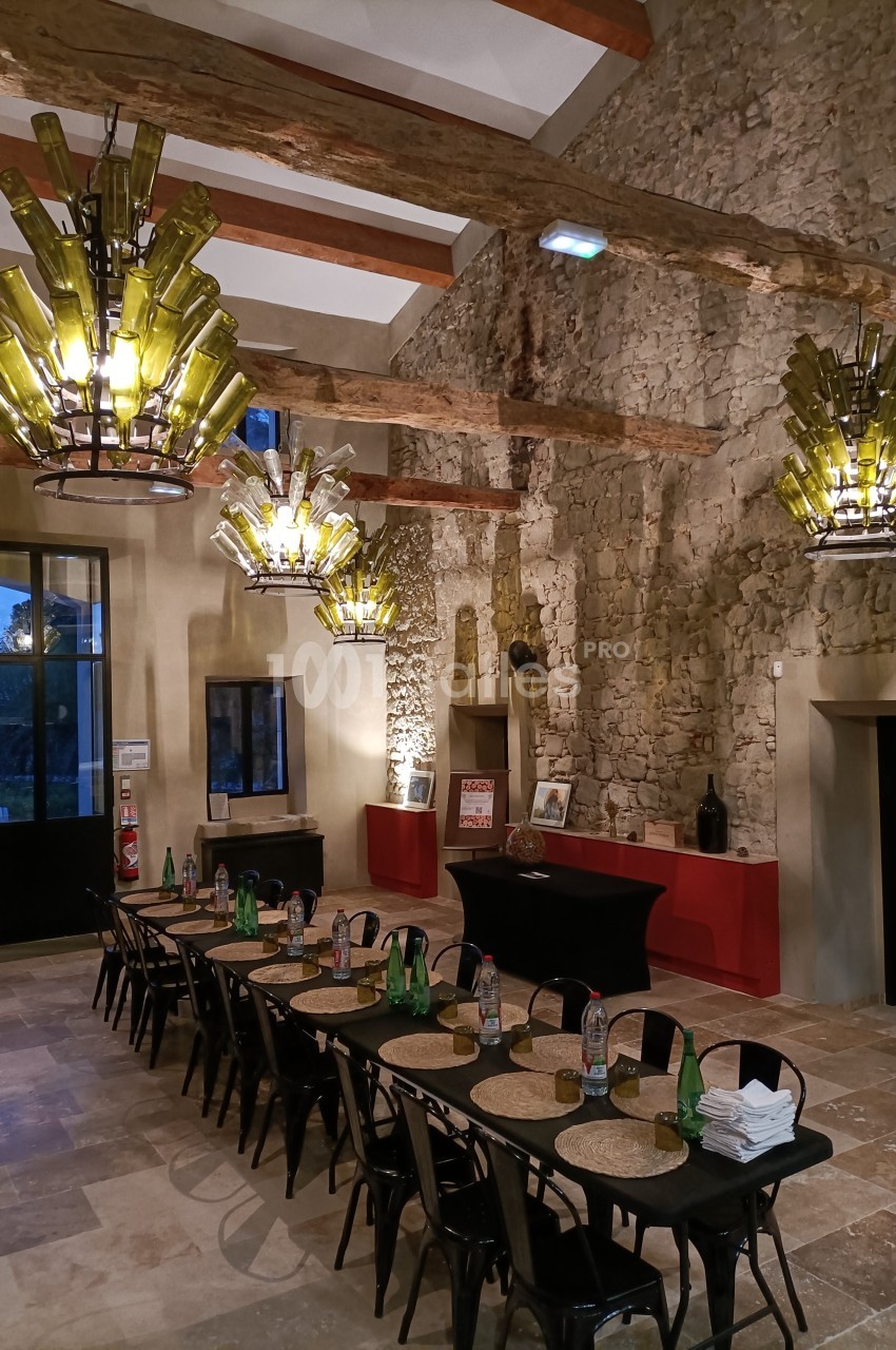 Salle en pierre avec de grandes tables dressées, éclairée par des lustres faits de bouteilles en verre vert.
