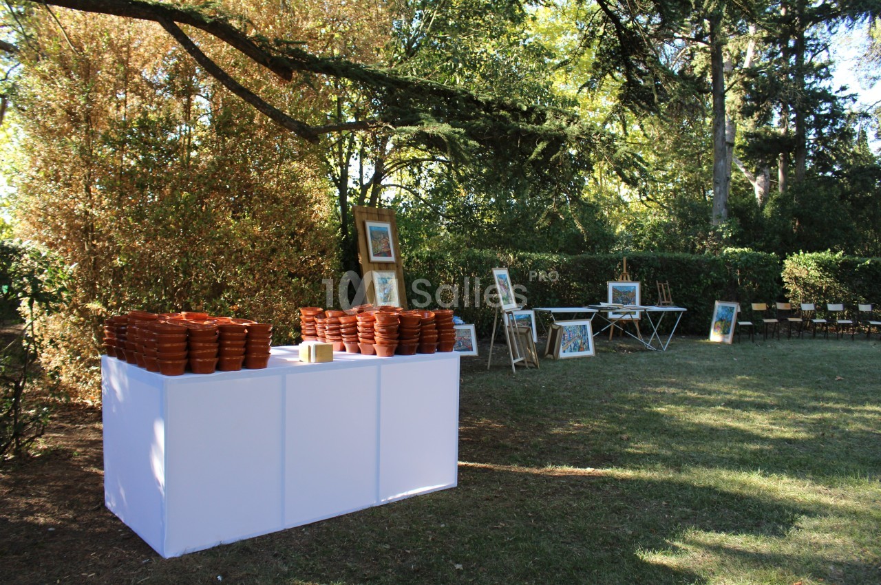 Table blanche avec des pots en terre cuite empilés, entourée de cadres et de chaises dans un jardin arboré.