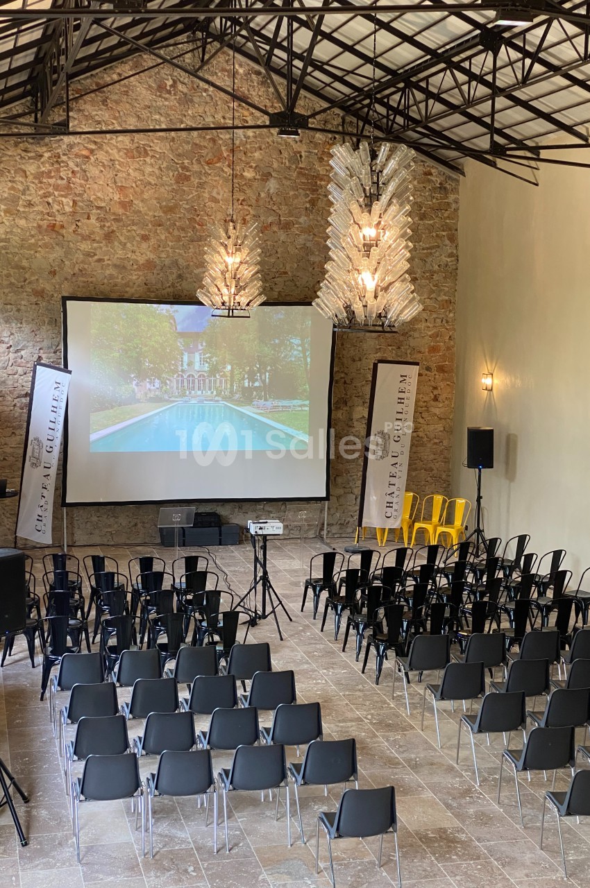 Salle de conférence avec des rangées de chaises, un écran de projection allumé et des bannières promotionnelles.