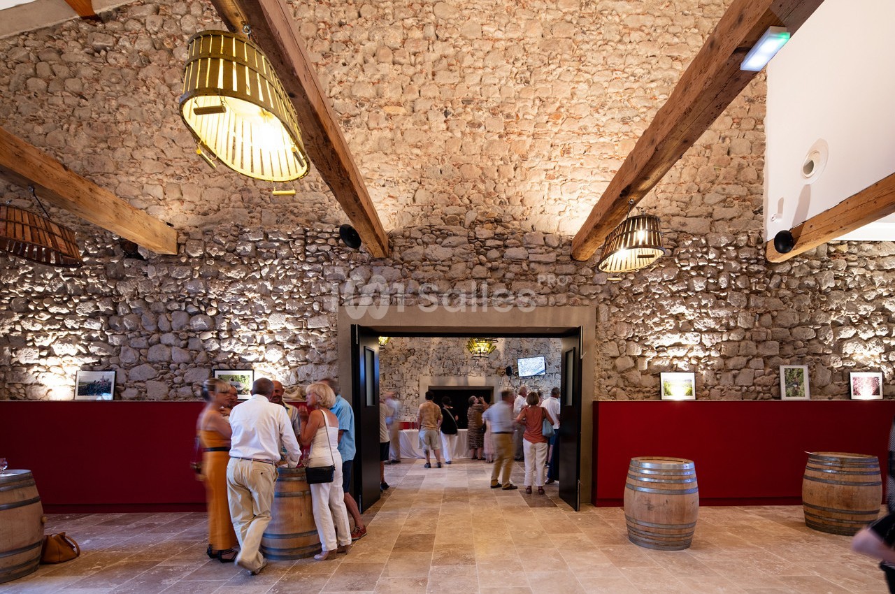 Salle en pierre avec poutres apparentes, visiteurs debout près de tonneaux et comptoirs rouges, éclairage chaleureux.