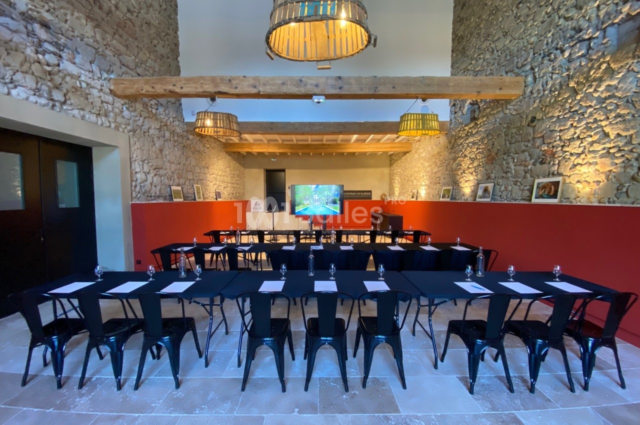 Salle de réunion avec murs en pierre, tables disposées en U, chaises noires et écran de présentation au fond.