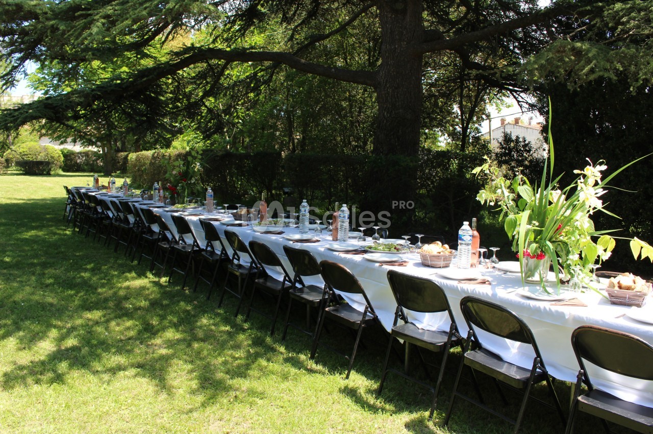 Table longue dressée en extérieur sous des arbres, avec nappes blanches, chaises noires et décoration florale.