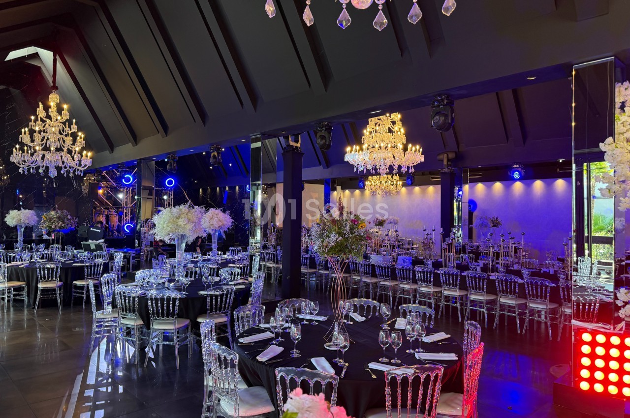 Salle de réception élégante avec tables dressées, chaises transparentes, lustres en cristal et éclairage d'ambiance.