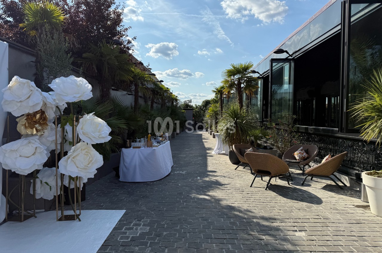Allée pavée bordée de plantes, avec tables décorées et fauteuils en osier, sous un ciel ensoleillé.
