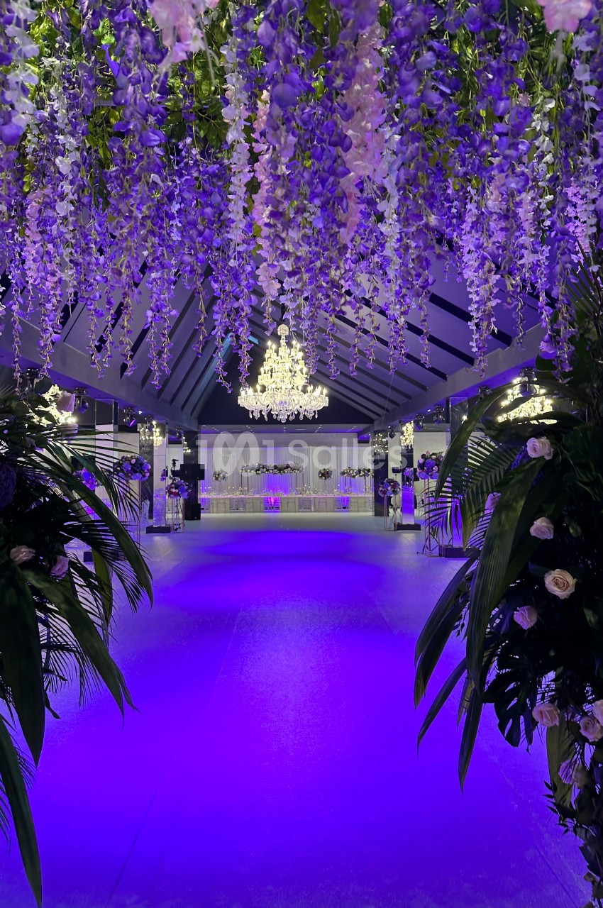 Salle de réception illuminée en violet, décorée de guirlandes florales suspendues et d'un grand lustre central.