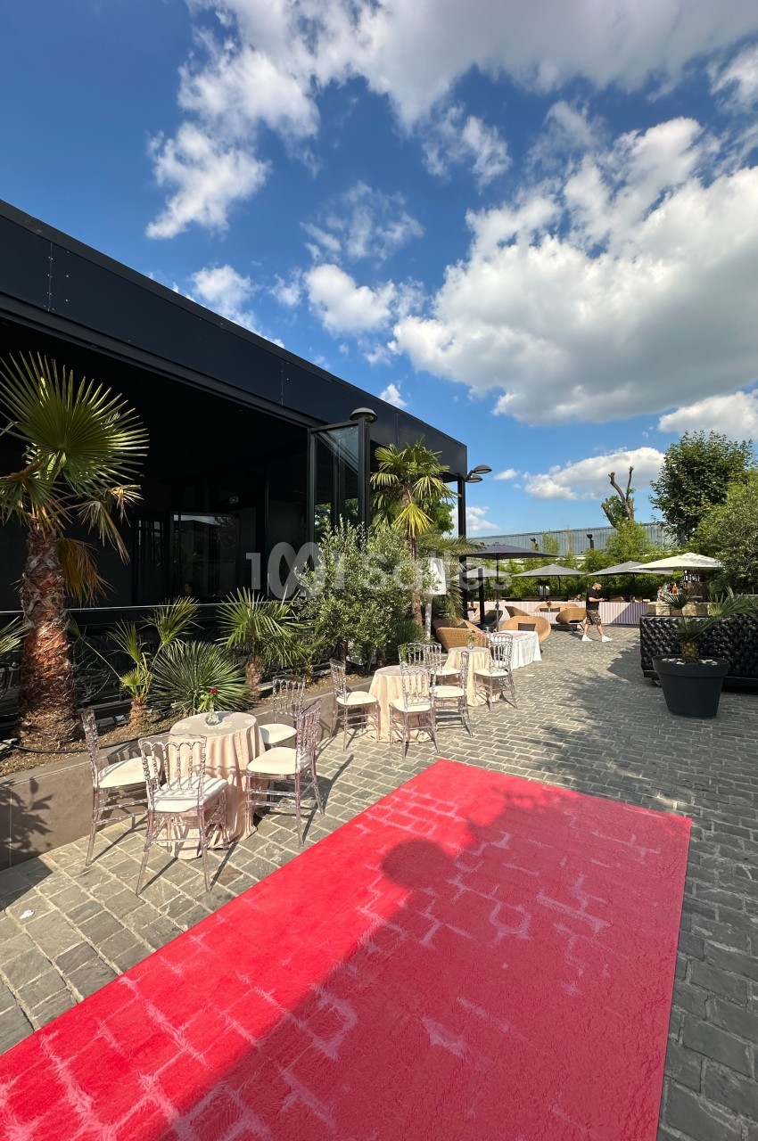 Terrasse extérieure avec tables et chaises, entourée de plantes et palmiers, sous un ciel partiellement nuageux.