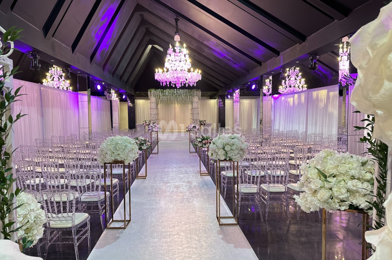 Salle de réception décorée pour un mariage, avec chaises transparentes, fleurs blanches et lustres suspendus.