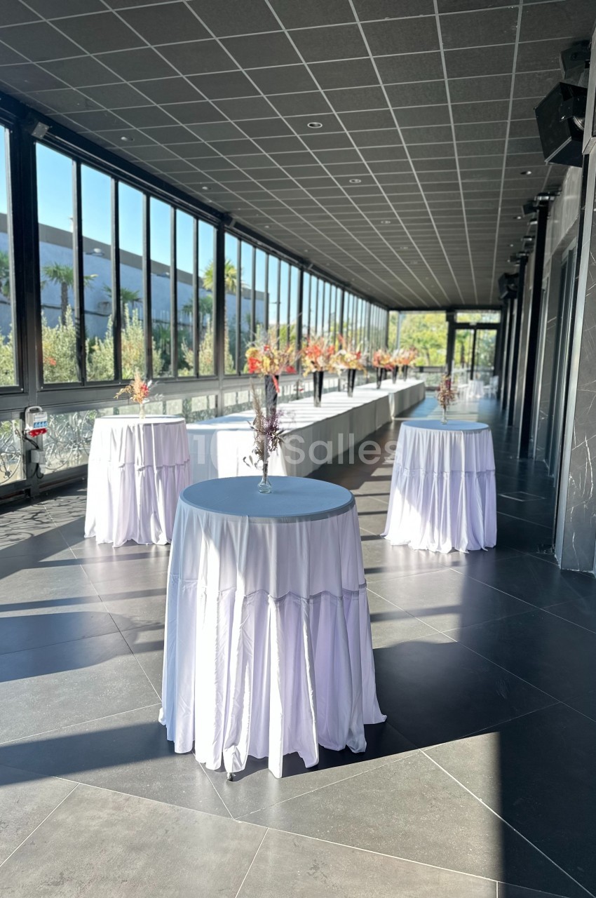 Salle lumineuse avec grandes baies vitrées, tables hautes drapées de blanc et décorations florales.