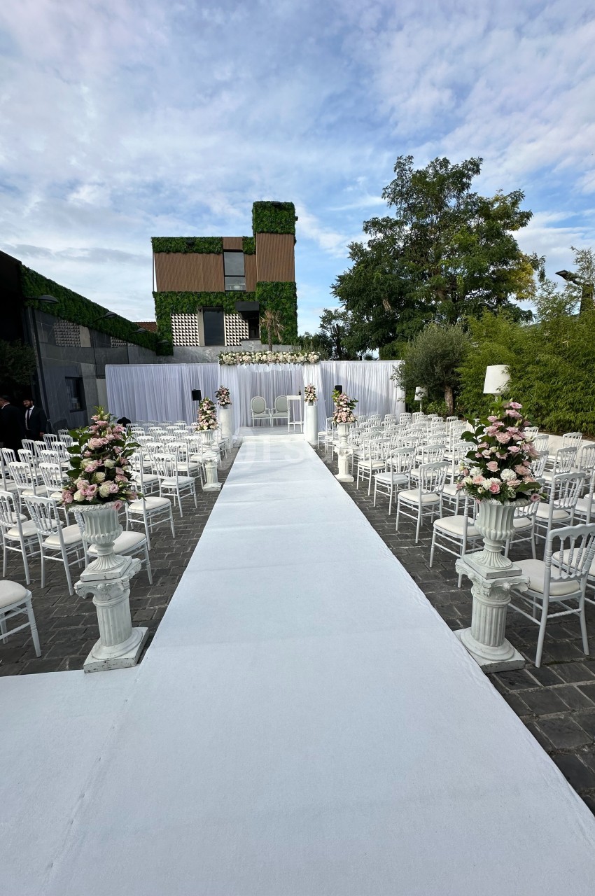 Allée centrale blanche bordée de chaises et de fleurs menant à un autel décoré pour une cérémonie en extérieur.