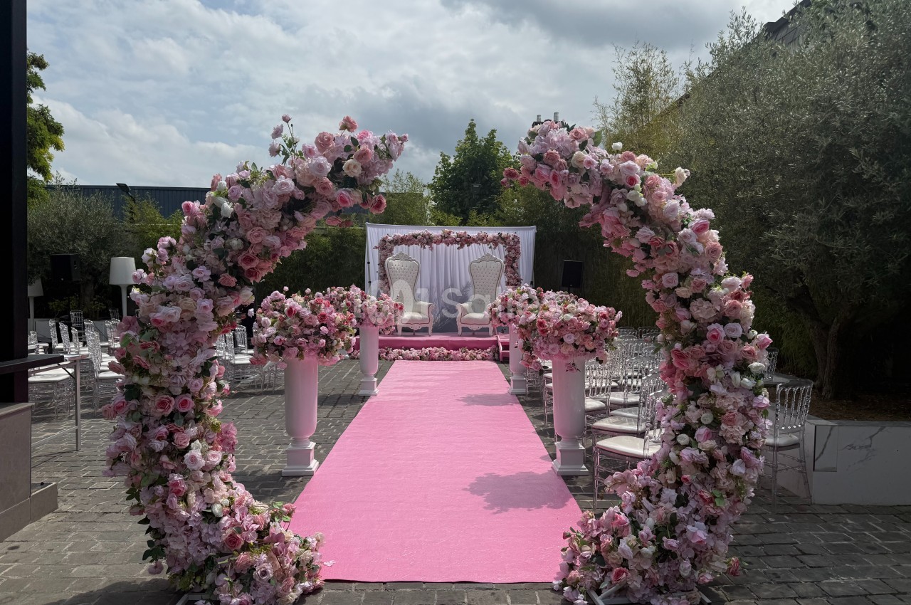 Allée centrale décorée de fleurs roses menant à une arche florale et deux fauteuils devant un fond blanc.