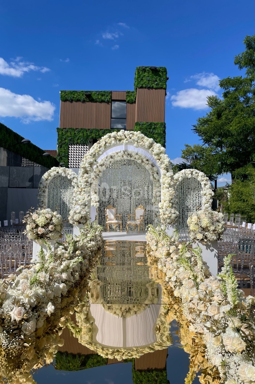 Allée décorée de fleurs blanches menant à une arche florale et deux chaises, avec un bâtiment moderne en arrière-plan.