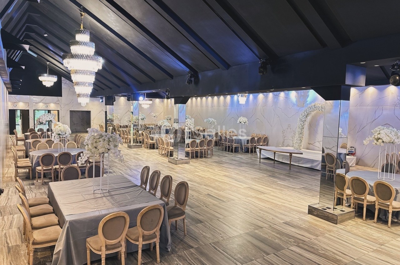 Salle de réception élégante avec tables dressées, chaises en bois clair, lustres suspendus et décorations florales blanches.