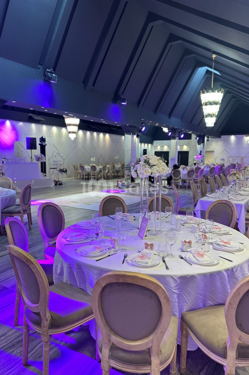 Salle de réception élégante avec tables rondes dressées, chaises en bois, éclairage violet et décorations florales.