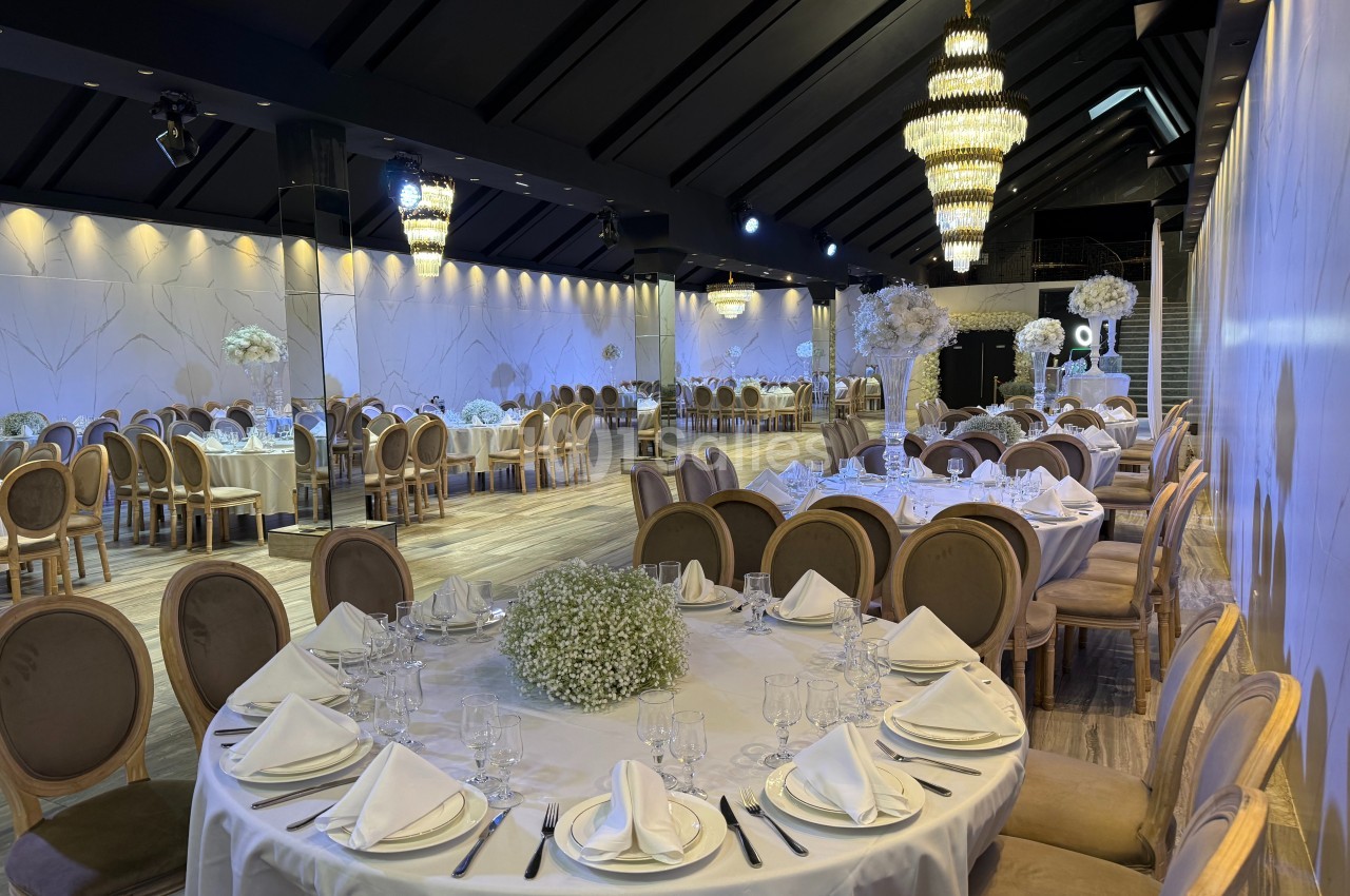 Salle de réception élégante avec tables rondes dressées, chaises en bois, décor floral blanc et grands lustres.