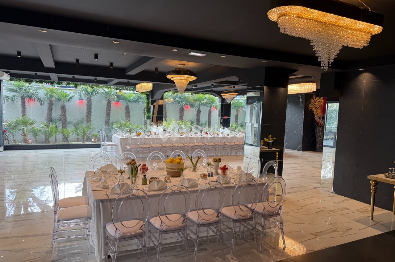 Salle de réception élégante avec tables dressées, chaises transparentes et lustres, donnant sur un jardin avec palmiers.