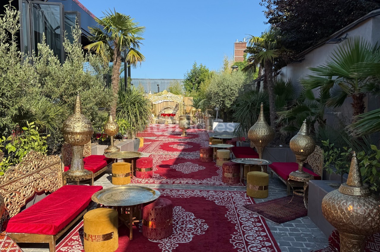 Allée décorée de tapis rouges, mobilier oriental doré et végétation luxuriante dans un cadre extérieur ensoleillé.