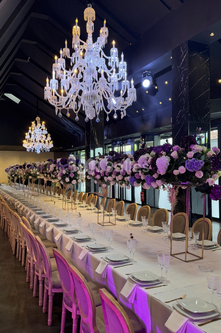 Salle de réception élégante avec longues tables dressées, chandeliers en cristal et décorations florales violettes.