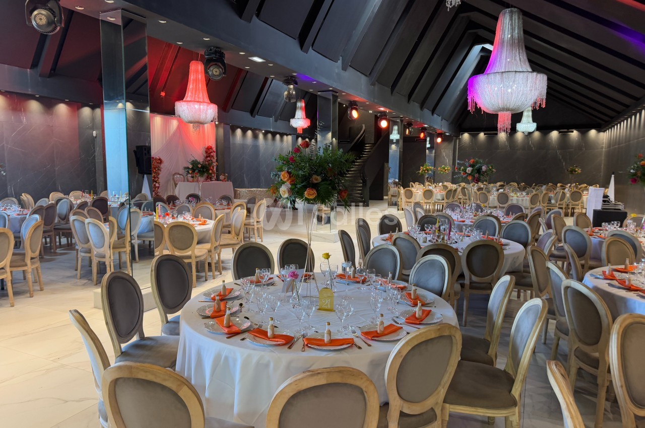 Salle de réception élégante avec tables rondes dressées, chaises en bois clair et lustres suspendus.