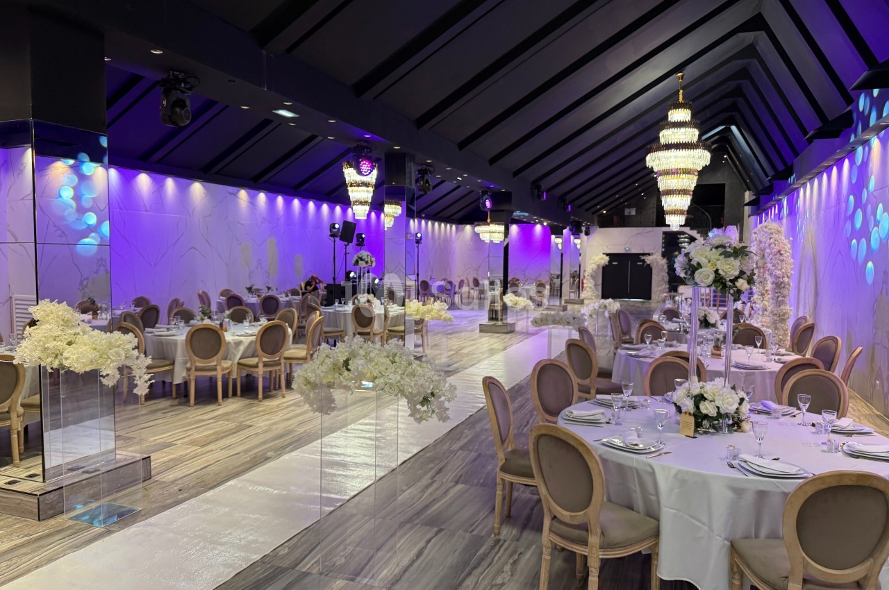 Salle de réception élégante avec tables dressées, décor floral blanc et lustres imposants sous un éclairage violet.